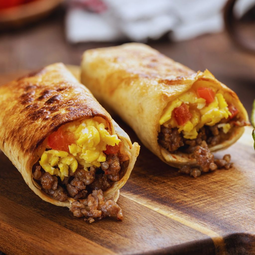 two breakfast burritos with eggs, red pepper, and sausage on a cutting board.