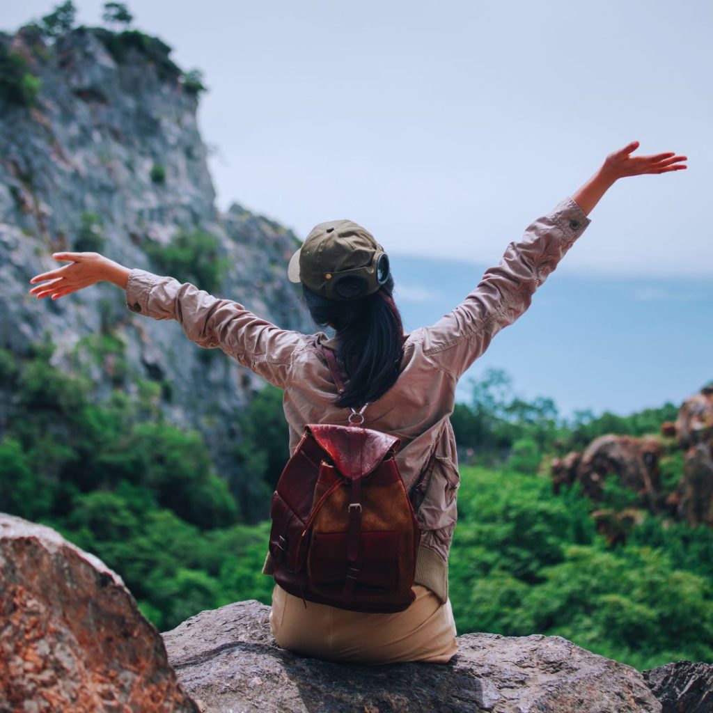 woman with a leather backpack sitting on a rock overlooking trees and water with her hands in the air